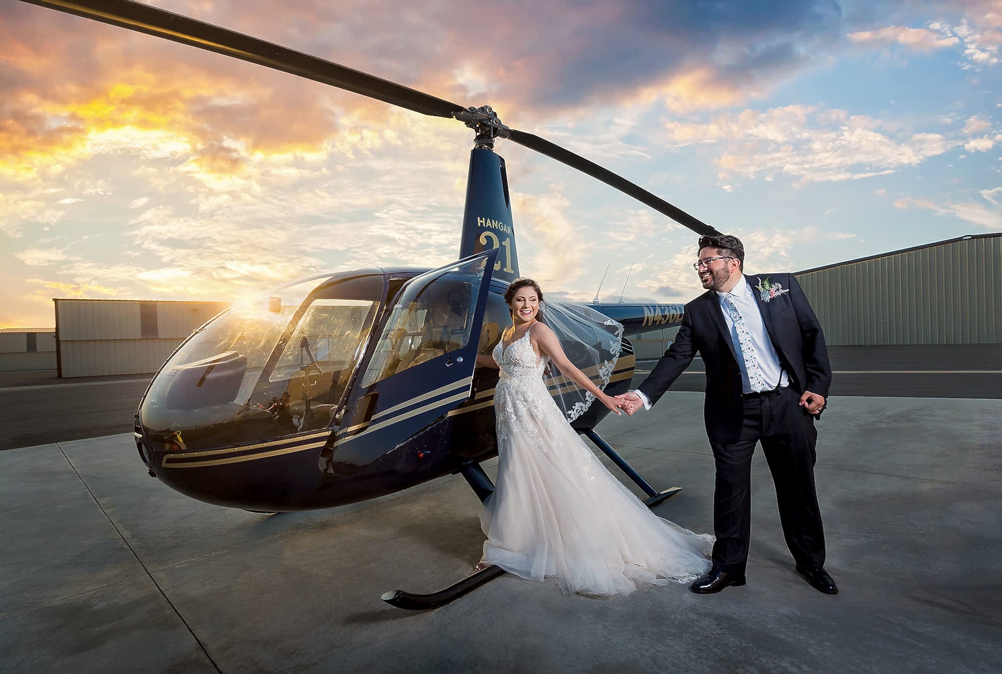 bride walking onto a helicopter outside of a hangar with a sunset in the distance while her groom holds her hand leading to grand entrance