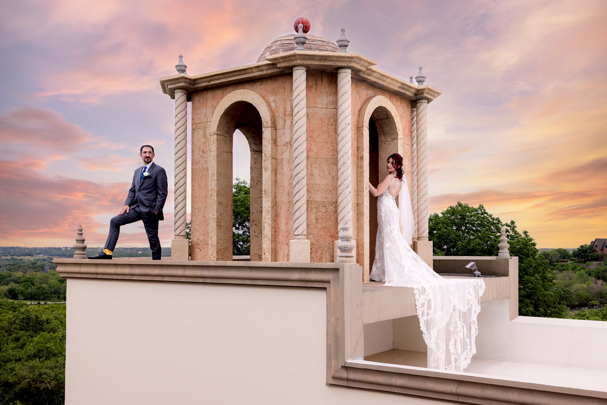 Bride and groom standing seperately on bell tower on Stoney Ridge Villa wedding venue's rooftop in Azle TX with intricate and laced wedding train flowing during cotton candy sunset behind them